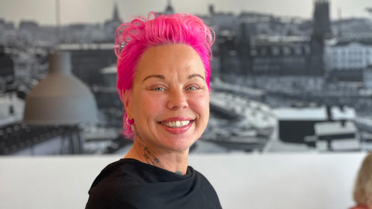 Glad projektledare för e-learning i rosa hår, står framför svartvit fototavla och håller i en mobil.