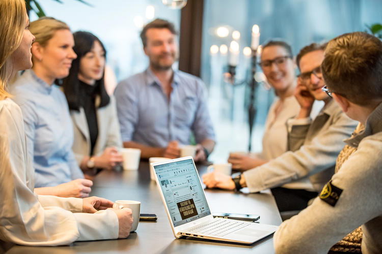 Grupp människor står runt ett loungebord med en dator på bordet.