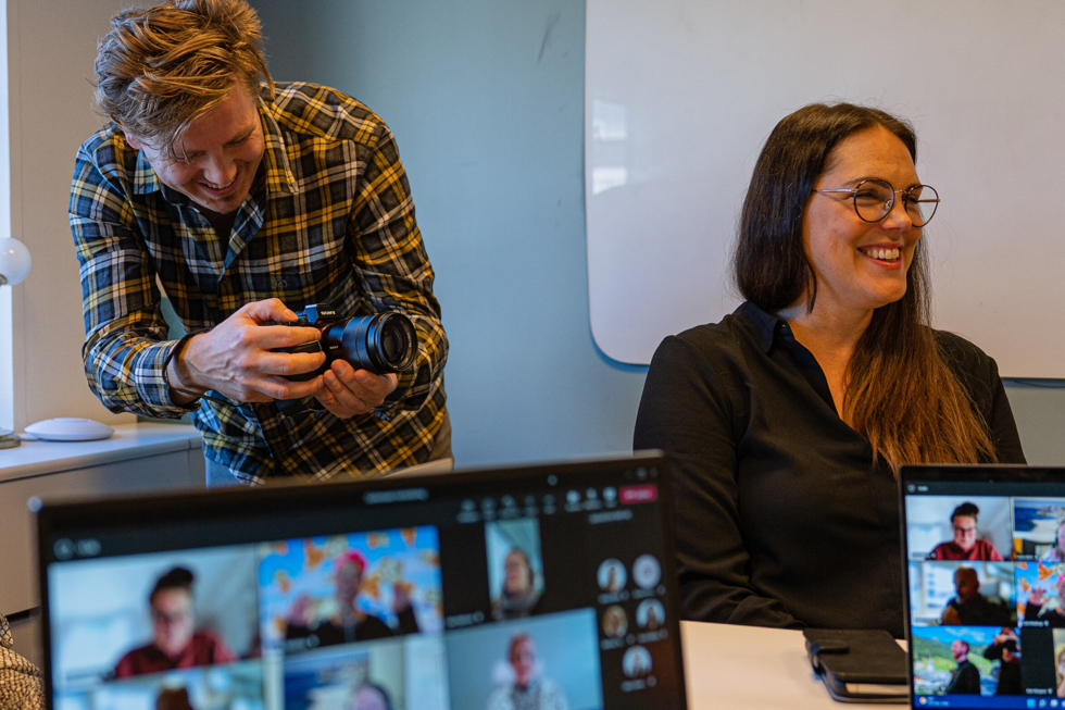 Flera skärmar med Teams-möten, i rummet fotar en kille och en tjej skrattar