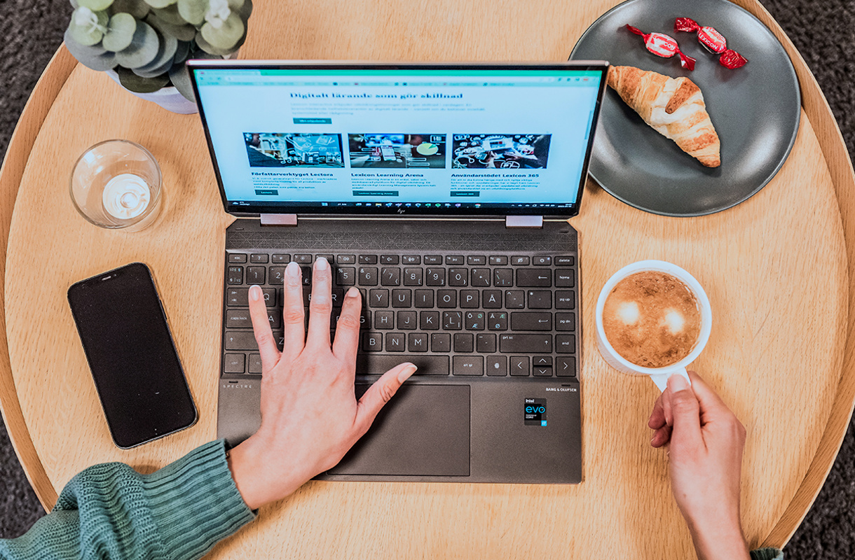 Vi ser uppifrån en hand på en laptops tangentbord och den andra handen som håller i en kaffekopp.