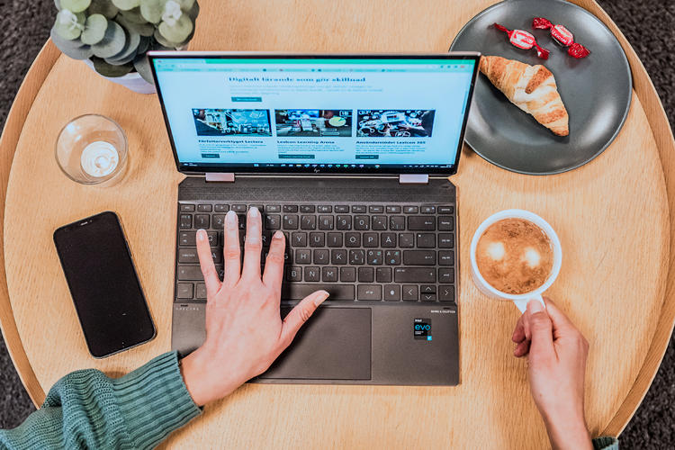 Vi ser uppifrån en hand på en laptops tangentbord och den andra handen som håller i en kaffekopp.